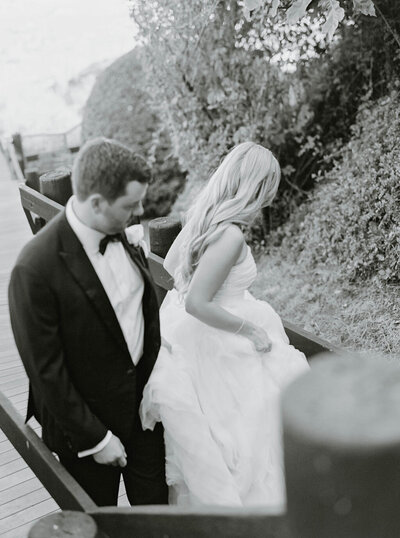 Rhode Island Wedding Photographer | A black-and-white photo of a bride and groom walking down a wooden path. The bride holds her dress, and the groom is in a tuxedo. 