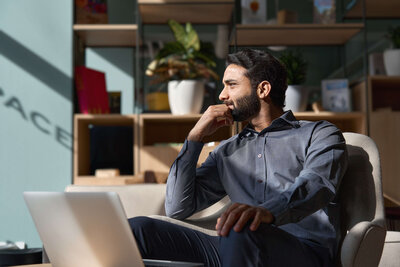 Thoughtful professional reflecting on strategy while seated with laptop in modern workspace.