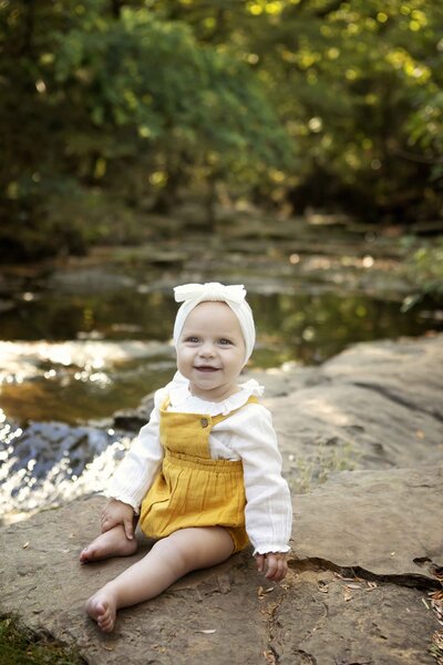 Birthday girl sitting happily by a creek, wearing a playful outfit, with the natural scenery and flowing water in the background