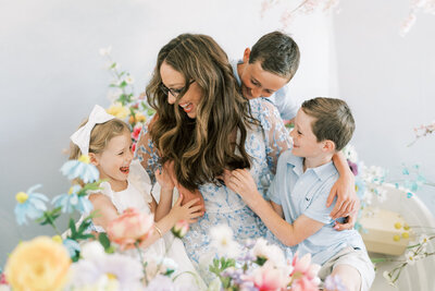 Mother having a tickle fight with her children in a photo studio filled with flowers.