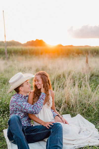 A couple sits on a blanket in an open field smiling at one another.