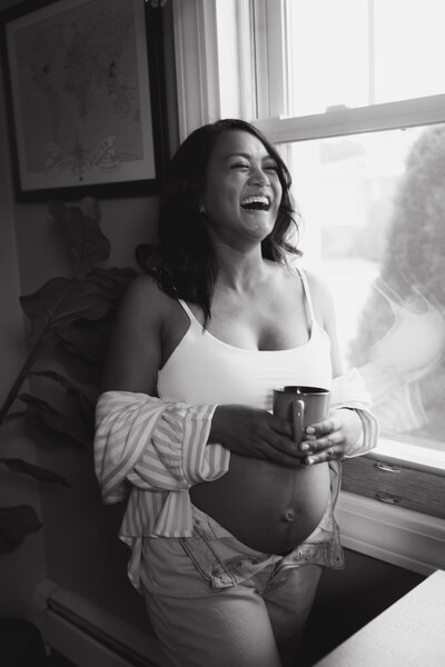 Pregnant woman laughs and holds a cup of coffee in her kitchen for maternity photos
