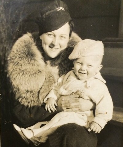 A 1935 photo of a young woman in a thick fur-collared coat and felt fascinator holding a  boy toddler on her lap wearing a white fleece jumpsuit.