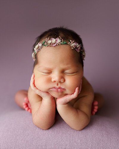 photo of newborn baby posed in a sitting position with hands propped on her wrists (froggy pose).  Baby has on lavender floral crown and is posed on a lavender fabric.