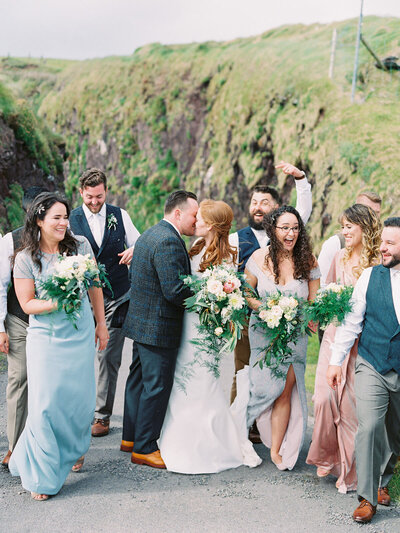 Wedding Photography in Dingle Ireland of a bride and groom kissing while their wedding party walks around them laughing wearing mismatched bridesmaid dresses