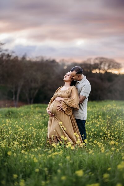 pregnant mama in mustard colored maternity dress leans calmly back on her husband. they stand in a dreamy wildflower field in lafayette, ca.