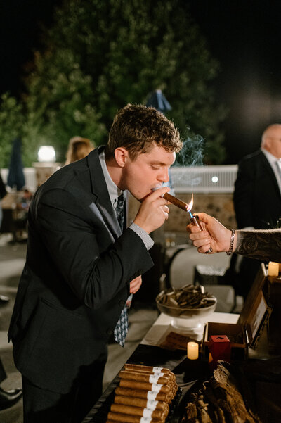 A wedding guest in a black suit leans forward to light a cigar at an outdoor cigar lounge during a wedding reception. He holds the cigar between his fingers while the cigar vendor extends a torch lighter, igniting the tip with a bright flame. Smoke curls into the night air as the guest takes the first puff. In the background, other wedding attendees are engaged in conversation, and string lights and candles add a warm glow to the evening atmosphere. A neatly arranged display of cigars rests on the table, inviting guests to partake in the experience. The setting captures the relaxed and celebratory ambiance of the wedding, where guests enjoy thoughtful details like a curated cigar bar under the stars.