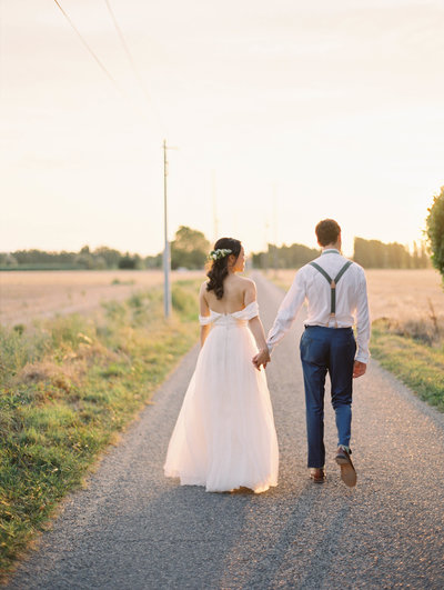 A bride and groom walk down a small strip of road.