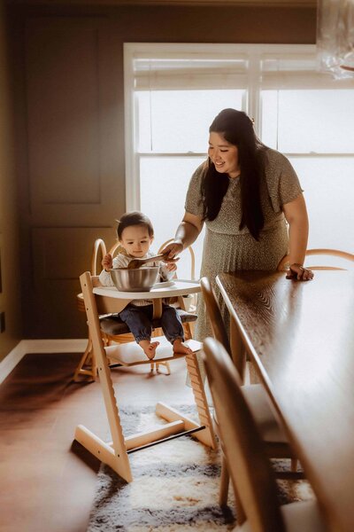 A woman stands beside a child seated in a high chair with a large metal bowl, inside a dining room filled with wooden furniture and natural light—a candid moment from a Warner Robins in home family photo session by Fire Family Photography.