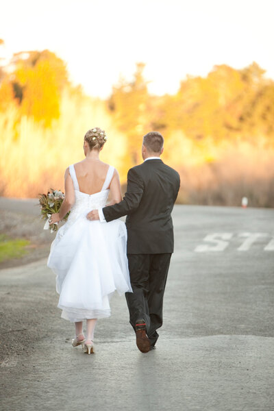 Eric and Phillipa Leseberg in wedding attire walk hand in hand along a quiet road, on their wedding day, with the bride holding a bouquet.