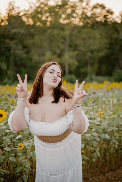 Senior girl in white giving kissy face and two peace signs in sunflower field in Maryland.