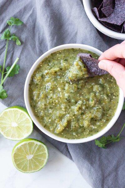 A hand scooping a tortilla chip into a bowl of green salsa.