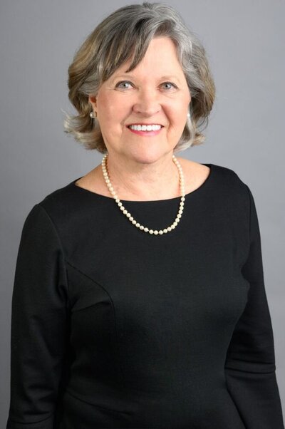 Smiling senior woman wearing a black dress, pearl necklace, and pearl earrings, posed against a plain gray background for a professional portrait.