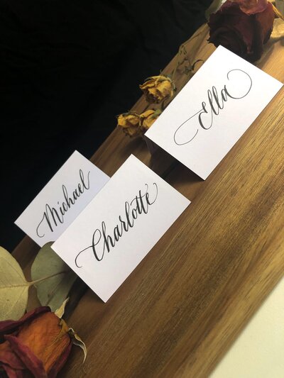 set of three calligraphy place cards against a black background and dried roses intertwined