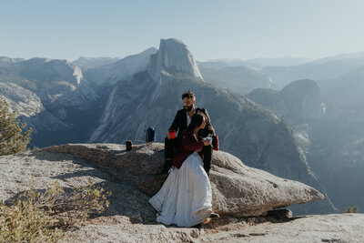 bride and groom sitting on rock together with Half Dome in the background they are holding coffee cups with their camping coffee maker next to them on the rock the bride is wearing a red down jacket over her wedding dress