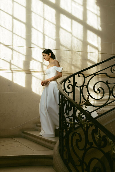 A bride walks down the staircase of a historic chateau, lit by a sunny window.