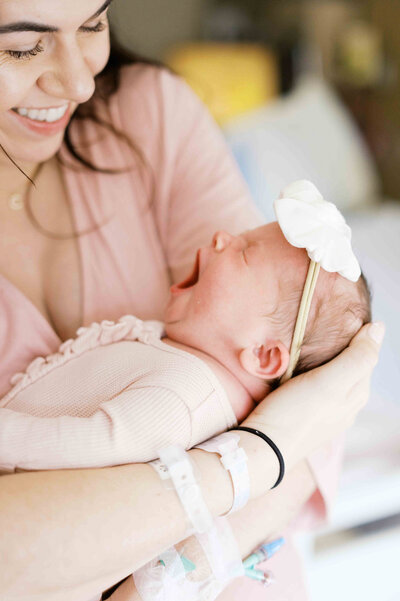 Mother wearing pink robe and hospital bracelets smiling down looking at newborn baby girl in pink ruffled pajamas with hair bow  by Portland Family Photographer Emilie Phillipson Photography. 