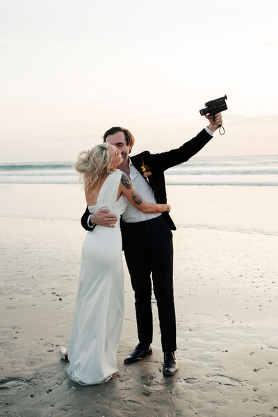 Bride and groom on film photography kissing and holding up a super 8 camera while on the beach after their ceremony on Anna Maria Island, Florida.
