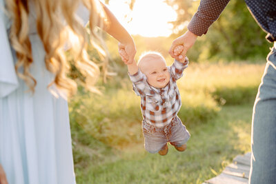 A joyful baby in a plaid shirt and jeans is held up by two adults' hands, swinging between them. Sunlight filters through the trees in the background, illuminating a grassy field—captured beautifully by Amber Bauer Photography, showcasing her knack for unforgettable family portraits.