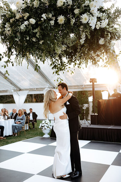 Lilly and Hunter share their first dance in the outdoor tent at Boxwood Estate, surrounded by soft lighting and elegant decor. The tent’s open sides offer views of the lush surroundings, while string lights and chandeliers create a warm, romantic ambiance. Lilly wears a beautiful white wedding gown, and Hunter is dressed in a classic suit. The couple smiles at each other, embracing the intimate moment in this picturesque outdoor setting.