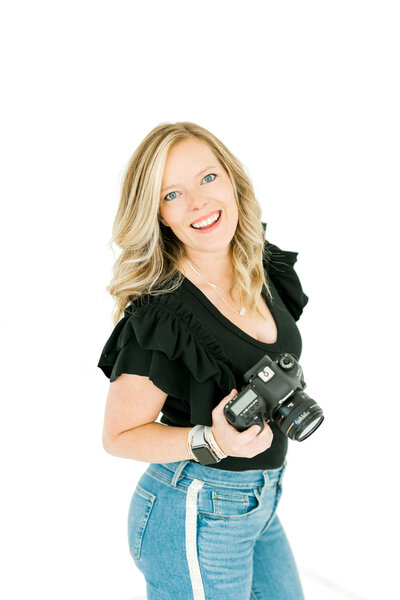 Brand photography kyle is holding her professional camera in her right hand. she is in front of a white wall and is wearing black blouse and jeans