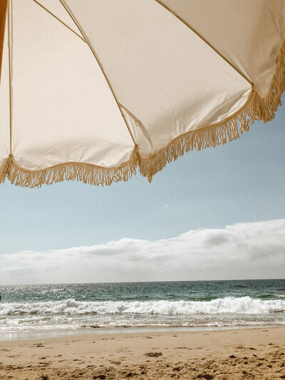 Let's Picnic Co.-Cream fringe umbrella overlooking the ocean at the beach