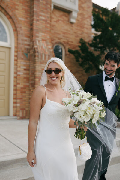 bride smiling wearing sunglasses and holding bouquet after exiting wedding ceremony 