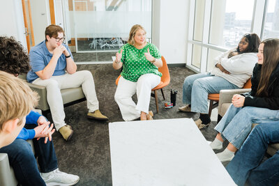 Six students sit in a circle around Mya Andersen, having a discussion on academic life coaching in a bright, modern room with large windows.