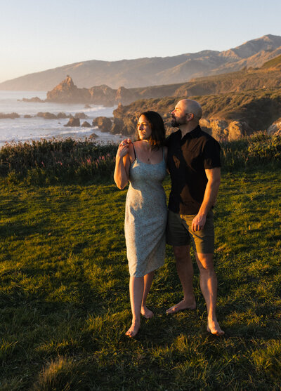 Becky & Brian, elopement photographers, stand side by side as the sunsets over the ocean at Big Sur, California.
