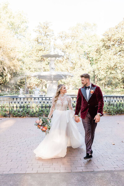 Bride and groom walking hand-in-hand along a brick sidewalk in Savannah, GA, surrounded by historic charm and greenery.