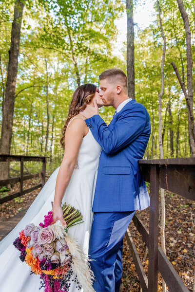 A bride and groom stand in a forest as he brings her in for a kiss. She is holding her bouquet. It is fall and the trees are yellow. Taken by Lexington Wedding Photographer.