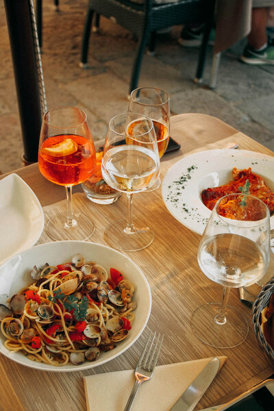 A table perfectly set after a day with your European travel specialist: one plate boasts spaghetti and clams, the other a savory meat dish. Four glasses—two with orange cocktails, two clear—await. Napkins and cutlery rest neatly on the wooden tabletop, ready for an exquisite dining experience.