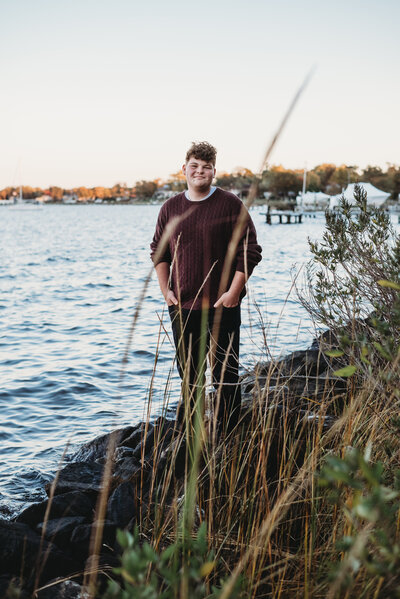 Senior photo of teen boy standing on rocks at waterfront in red sweater and black pants in Annapolis Maryland.