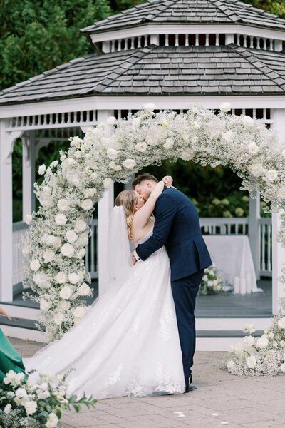 Groom kisses bride in front of round white floral arch at outdoor ceremony at Deer Creek, photographed by a professional wedding photographer in Toronto