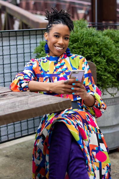 black woman sitting outside at table in colorful dress holding cell phone