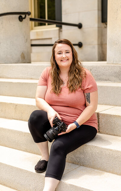 Woman with brown hair sitting on stairs wither her camera in her hand . She is smiling and wearing a coral colored t-shirt and you can see a tattoo on her arm