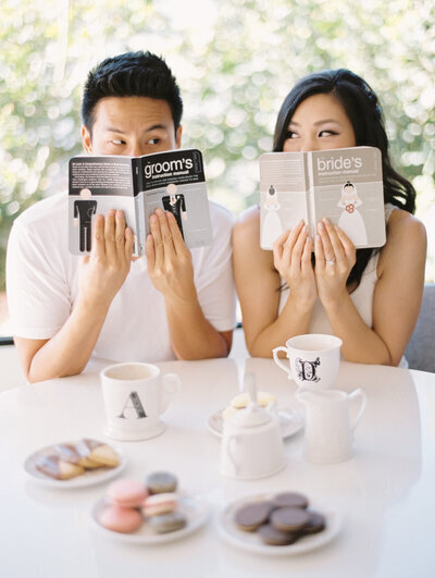 Kohnur Bride with soft glam makeup and romantic loose curls shares a playful coffee date moment with her groom, both smiling behind matching bride and groom books in a cozy engagement shoot