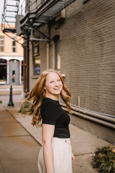 Smiling young woman in a black top and beige pants walking in an urban alleyway.