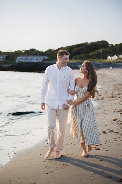 Engagement Photos Rhode Island | Kelsey Sheehan Photography Timeless Rhode Island Weddings | A couple walks hand in hand on a sandy beach at sunset, smiling warmly at each other. The ocean and distant houses provide a serene backdrop.