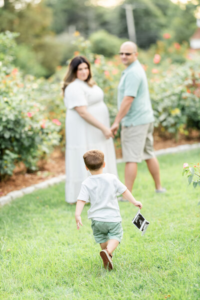 An expecting mother wearing a beautiful pink dress is standing between green shrubs as she gently caresses her bump photographed by Milford CT Photographer, Tambella Photography