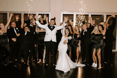 A bride and groom strike a celebratory pose with their wedding party in front of indoor sparklers at SoHo63, a Chandler wedding and event venue. The groom wears a white tux jacket, the bride beams in a fitted lace gown, and their guests cheer in matching black formalwear.