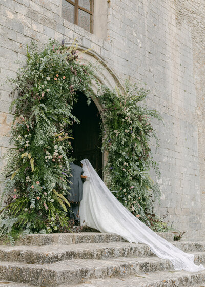 A bride's long veil trails behind her as she enters a mountaintop stone building in Sicily.