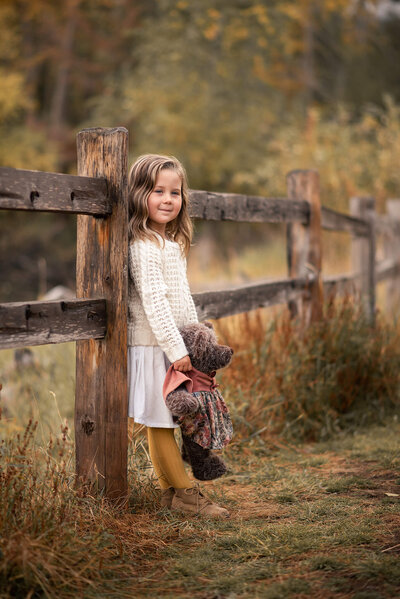 Little girl leaning against a wooden fence while holding a teddy bear, smiling at the camera. Portrait by Brittany Danielle Photography