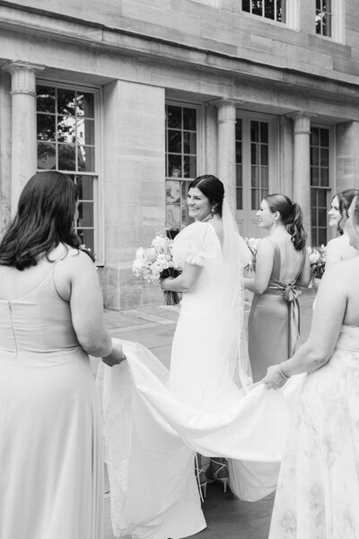Black and white image of bride walking while bridesmaids hold her train