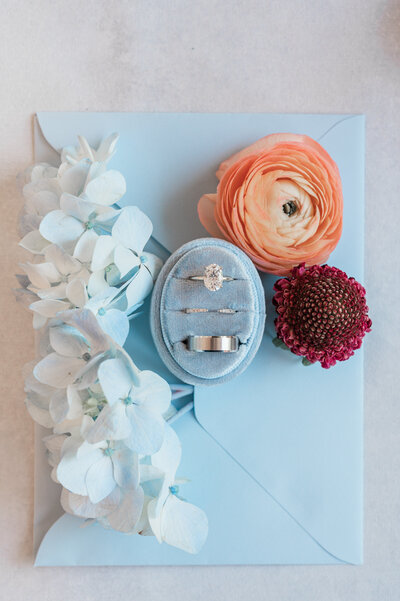 A blue ring box with three rings in it next to small flowers