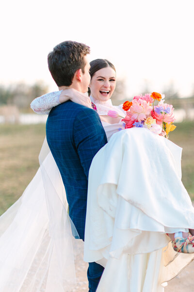 Groom carrying bride while she holds a colorful bouquet