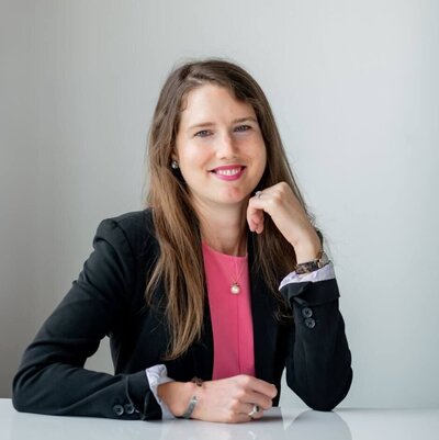 Brenna Clair Moore smiling in a black blazer and pink top, seated at a white desk with a light gray background.
