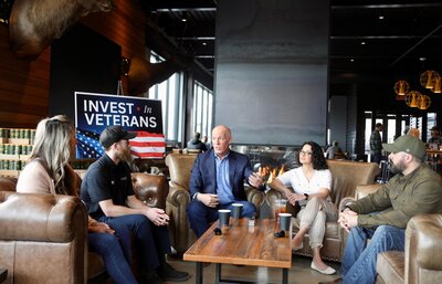 Montana Governor Greg Gianforte speaking with a panel of veteran-owned small business leaders during an Invest in Veterans event at a fireside roundtable in a modern lodge setting.