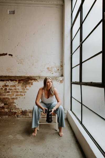 Kaz Soom, Perth Maternity, Newborn and Family Photographer.  She is sitting on a white folding chair, holding her camera and looking out the window. 