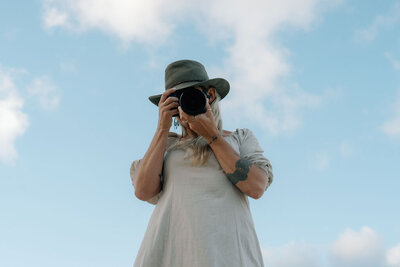 Woman wearing a wide-brim hat holding a camera up to her face with a bright blue sky behind her
